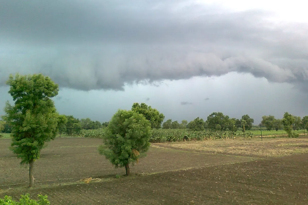 A dark shelf of monsoon rain clouds rolling over ploughed brown fields and scattered trees in a village in Maharashtra