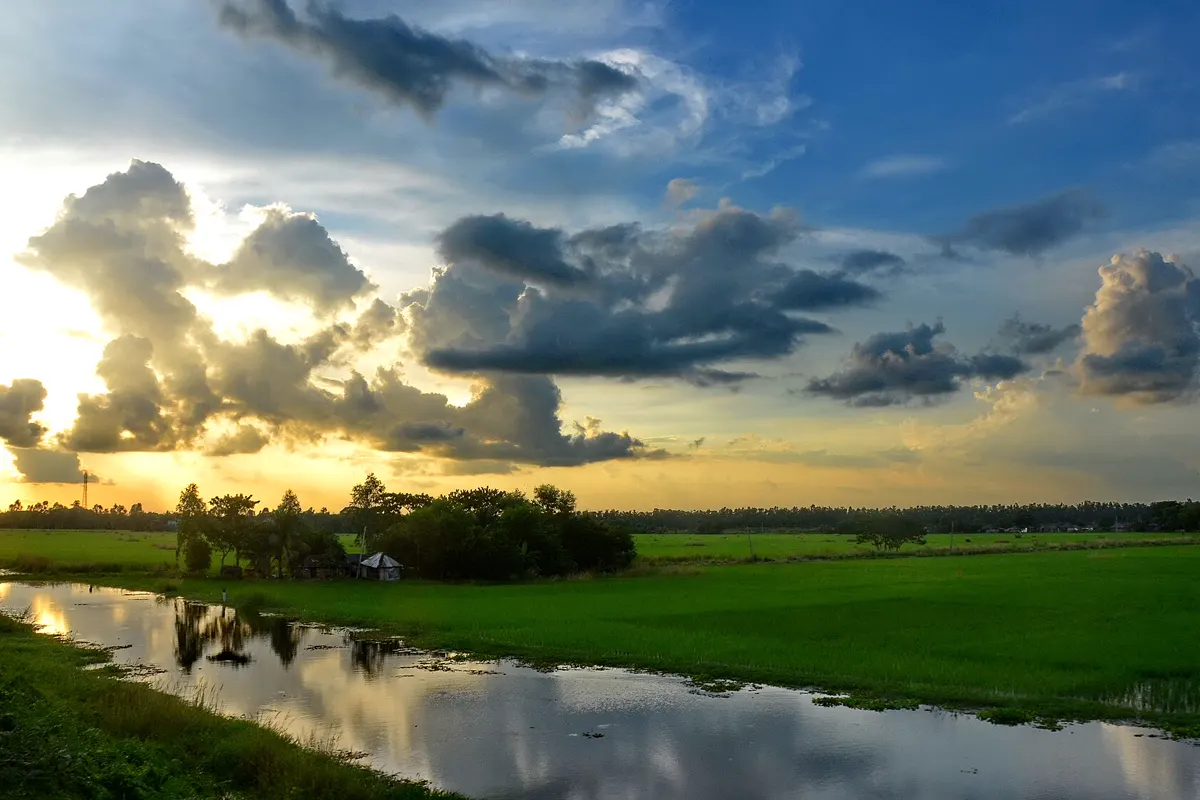 Monsoon clouds lit by a setting sun over flat green fields and a still canal in rural West Bengal