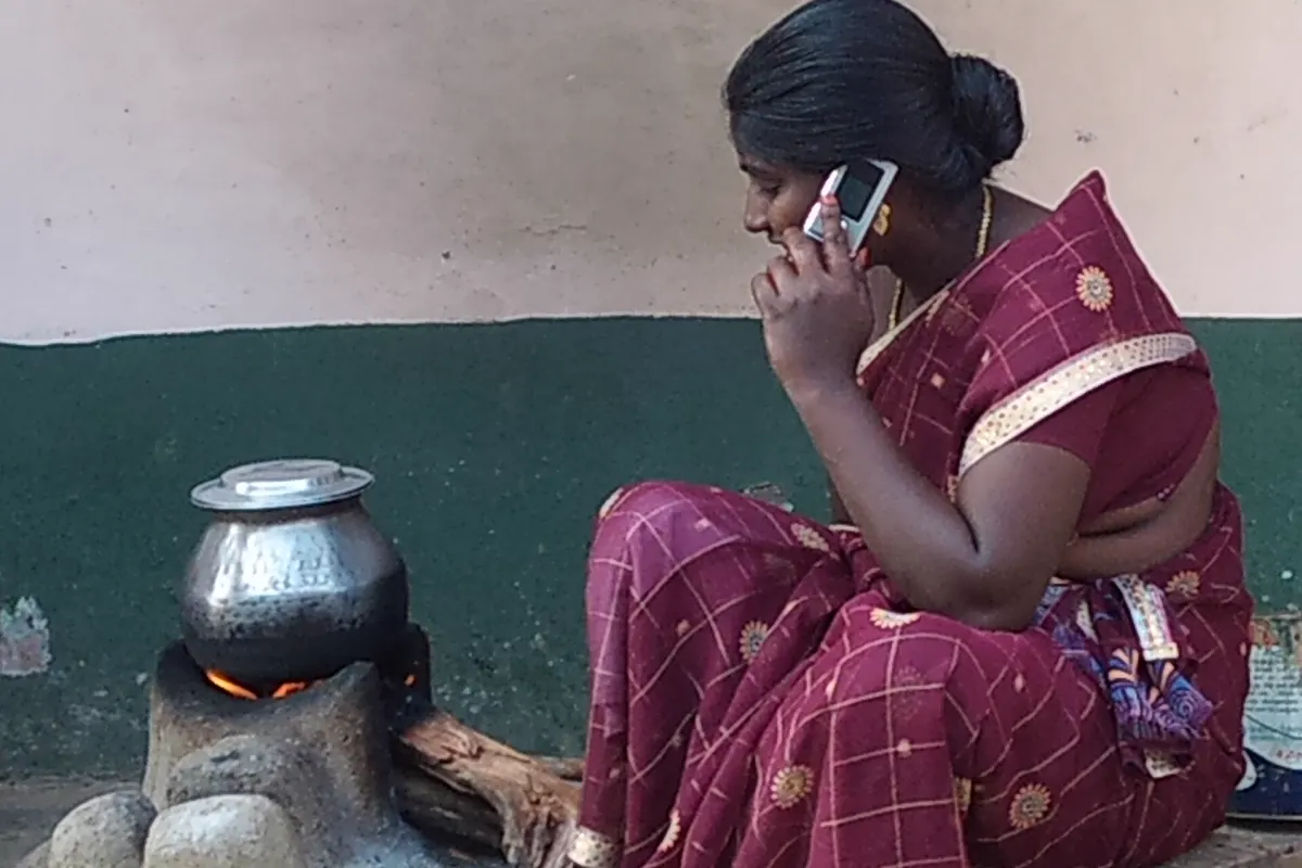 A woman in a maroon sari talking on a mobile phone while sitting beside a metal pot on a wood-fired stove in rural Tamil Nadu