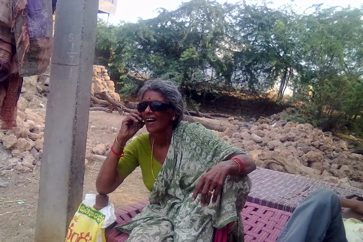 An older woman in a green sari and sunglasses smiling as she talks on a mobile phone outdoors in a village in Andhra Pradesh